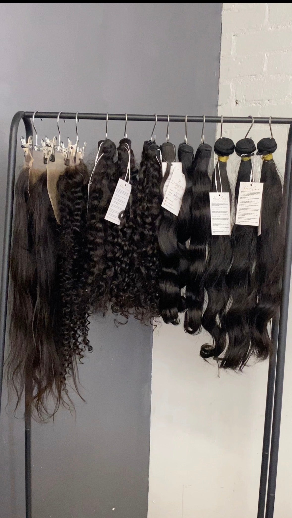 Hair extensions hanging on a rack with labels against a gray wall.